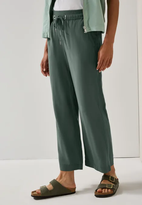 Person trägt grüne Hose und Sandalen, stehend vor weißem Hintergrund.